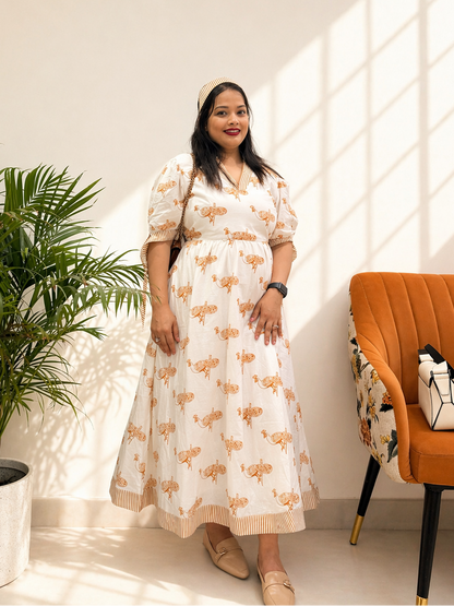 Woman in a white dress with orange patterns standing in a bright room with a plant and chair.