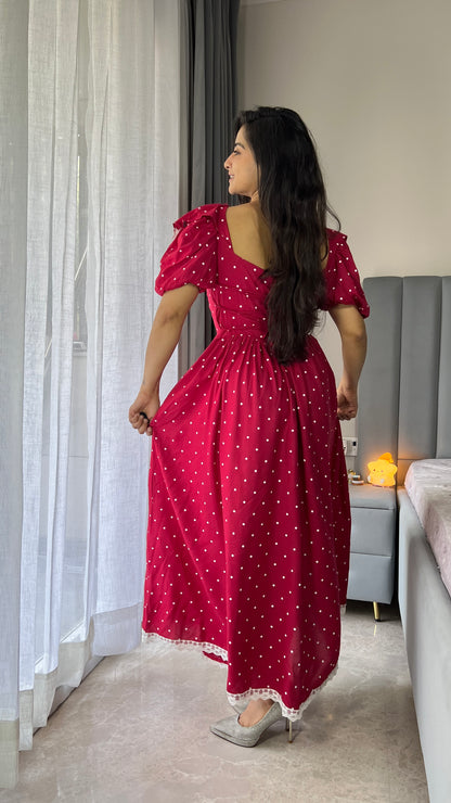 Woman in a red polka dot dress standing in a room with curtains and a sofa.